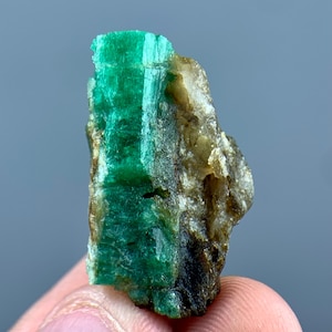 Puede incluir: Un espécimen de cristal de esmeralda en bruto que presenta una columna hexagonal verde vibrante y translúcida integrada en una matriz rugosa de color marrón tierra y gris. El mineral muestra estrías verticales naturales y texturas cristalinas.