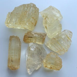 May include: A selection of raw topaz crystals in shades of pale yellow and gold. The uncut gemstones vary in shape and size, showcasing internal structures and textures. The crystals are presented on a white background.