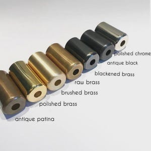 May include: A collection of cylindrical metal beads in various finishes, including polished chrome, antique black, and raw brass. The image displays the beads in a diagonal row, with text labels identifying each finish. The beads are approximately 1 inch in length.