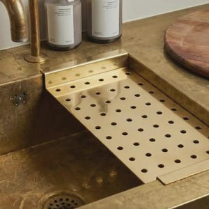 May include: A brass kitchen sink with a built-in perforated drainboard. The sink has a brass faucet and two bottles on the countertop. A wooden cutting board sits on the right side of the sink.