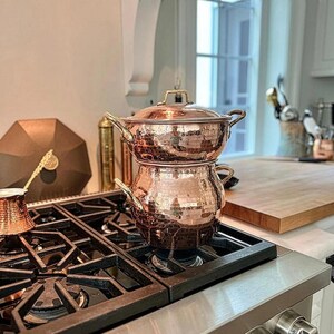 Pode incluir: Conjunto de panelas de cobre com acabamento martelado, incluindo uma panela com tampa e uma panela menor empilhada em cima. O conjunto está em um fogão a gás preto. Uma pequena cafeteira de cobre também é visível.