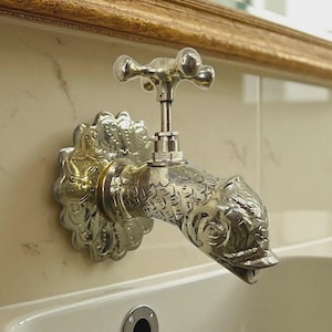 May include: Ornate silver-toned faucet with a fish-shaped spout. The faucet is mounted on a marble-patterned wall, with a decorative floral base and a cross-shaped handle. A gold-framed mirror is visible in the background.