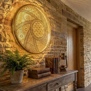 Peut inclure: Un luminaire mural circulaire avec un motif ajouré complexe est monté sur un mur en pierre. Une console en bois contient une fougère en pot, des livres et une lanterne. La pièce dégage une ambiance chaleureuse et rustique.