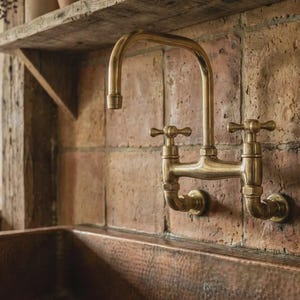 Puede incluir: Un grifo de cocina de latón de estilo vintage, montado en una pared de ladrillo. El grifo tiene dos manijas en forma de cruz y un caño curvo. Debajo se ve un fregadero de cobre. La escena evoca una estética rústica.