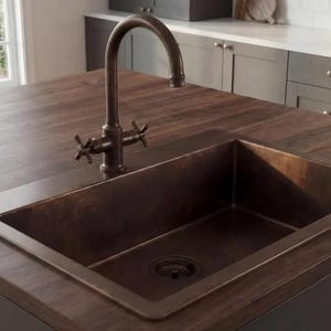 May include: A copper-colored kitchen sink with a matching faucet is set into a dark wood countertop. The sink is rectangular with a drain in the center. The faucet has a curved spout and cross handles. The kitchen island is dark brown.