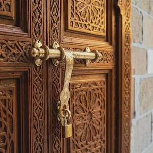 May include: Close-up of a wooden door with intricate carvings and a brass latch. The door is a warm brown color, and the carvings feature geometric and floral patterns. A brass lock secures the door.
