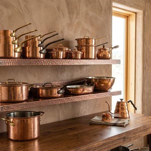 Puede incluir: Una colección de utensilios de cocina de cobre pulido que incluye varias ollas, sartenes y teteras dispuestas en dos estantes de pared de cobre martillado y una mesa de madera. El conjunto presenta asas metálicas, tapas y una tetera.
