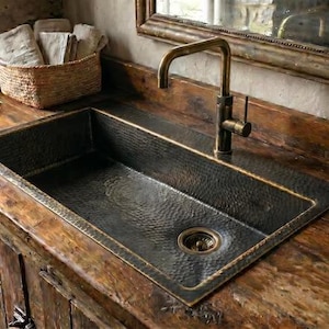 May include: A rectangular hammered dark metal sink set into a rustic wooden countertop. The sink features a central drain and is paired with a tall, curved, antique brass-toned faucet. The metal surface has a textured, dimpled finish throughout.