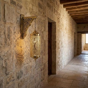 Puede incluir: Una lámpara de pared dorada y ornamentada, con una pantalla cilíndrica calada, cuelga de una pared de piedra. El intrincado diseño de la lámpara proyecta una luz estampada. El pasillo tiene suelo de piedra y techo de madera.