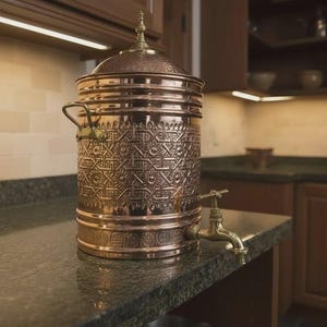 May include: A copper water dispenser with an ornate, engraved design. The dispenser has a spigot and a lid with a decorative finial. It is sitting on a countertop in a kitchen setting.