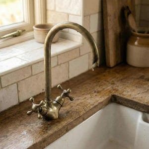 Op de afbeelding: Een messing kraan in vintage-stijl met kruisvormige handgrepen is gemonteerd op een houten aanrechtblad. De kraan heeft een gebogen tuit en er is een waterdruppel zichtbaar. De gootsteen is wit en de achtergrond omvat een raam en een kleine keramische pot.