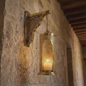 May include: A gold-colored, ornate wall-mounted lantern with a candle inside. The lantern hangs from a decorative bracket on a stone wall. The lantern's design features intricate cut-out patterns, casting light and shadow.