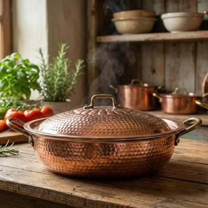 Puede incluir: Una olla de cobre con textura martillada y tapa a juego. La olla tiene dos asas y un asa en la tapa. Sale vapor de la olla, lo que sugiere que está en uso. Otros utensilios de cocina de cobre son visibles en el fondo.