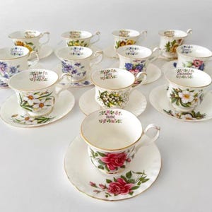 May include: A collection of vintage teacups and saucers, each adorned with floral designs and the name of a month. The teacups feature various flower patterns, including roses, daffodils, and lilies of the valley, with gold trim.