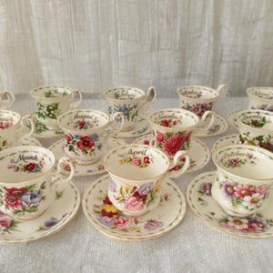 Könnte beinhalten: Eine Sammlung von Vintage-Tassen und Untertassen, jede mit floralen Mustern und dem Namen eines Monats verziert. Die Tassen haben zarte Henkel und stehen auf passenden Untertassen. Die floralen Designs umfassen rote, rosa und lila Blumen.