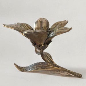 Op de afbeelding: Een bronzen, bloemvormige kandelaar. De kandelaar is ontworpen om op een bloem te lijken met gedetailleerde bloemblaadjes en bladeren. Het metaal heeft een licht verouderd uiterlijk, wat bijdraagt aan de vintage charme. Het ontwerp is ingewikkeld en elegant.