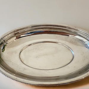 Può includere: Un vassoio da portata ovale color argento lucido con una superficie liscia e riflettente e un bordo rialzato con dettagli a perline. Il vassoio presenta un bordo largo e piatto che degrada verso un centro concavo e poco profondo.