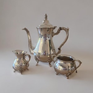 Puede incluir: Un juego de té plateado que incluye una tetera, una jarra para la crema y un azucarero. La tetera tiene un pico curvo y un asa ornamentada. La jarra para la crema y el azucarero tienen asas pequeñas y se apoyan en tres patas.
