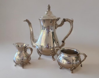 May include: A silver tea set including a teapot, creamer, and sugar bowl. The teapot has a curved spout and ornate handle. The creamer and sugar bowl have small handles and sit on three feet. The set is reflective and sits on a white surface.