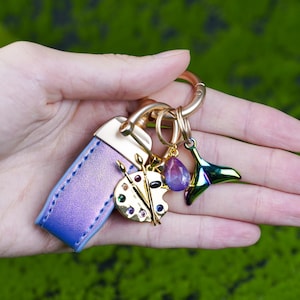 Peut inclure: Un porte-clés avec une lanière en cuir violet et des éléments dorés. Il comprend un charm en forme de palette d'artiste dorée avec des détails de peinture colorés, un charm en forme de goutte violette et un charm en forme de queue de baleine verte irisée.