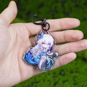 May include: A clear acrylic keychain featuring a detailed anime-style character with white hair and blue accents. The character is wearing a blue and white outfit with a peacock feather design. The keychain has a black metal clasp.