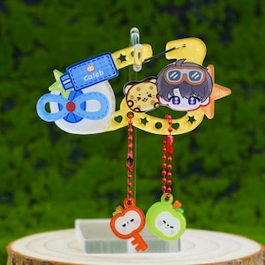 Könnte beinhalten: Ein gelber Acryl-Karabiner-Anhänger mit einer Cartoon-Figur mit Schutzbrille, einem kleinen Leopardenkopf und einer blauen Tube mit der Aufschrift Caleb. Zwei rote Kugelkettchen hängen daran, jeweils mit einem kleinen apfelförmigen Anhänger.