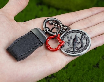 May include: A keychain featuring a black leather strap, a red accent, and a silver-coloured round charm. The charm has an engraved design with text around the edge. The keychain is held in a hand against a green background.