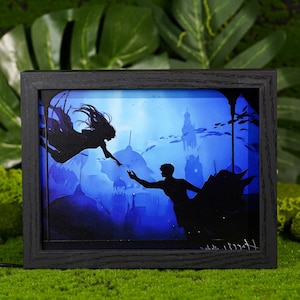 Peut inclure: Une œuvre d'art encadrée représentant deux silhouettes se tendant la main dans une scène sous-marine. L'œuvre présente un cadre en bois foncé et une palette de couleurs bleue et blanche, avec des poissons et des éléments architecturaux visibles.