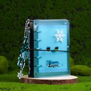 Può includere: Un raccoglitore blu trasparente con un motivo a fiocco di neve e il nome "Zayne" stampato sul davanti. Un ciondolo a fiocco di neve abbinato è appeso al raccoglitore. Il raccoglitore è fissato con un elastico nero.