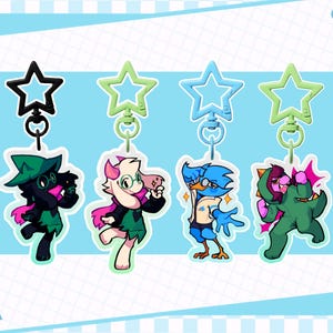 May include: Four character keychains with star-shaped toppers. The characters are in various colors, including black, pink, green, and blue. The background is light blue with a white border and a checkered pattern.