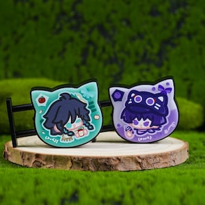 May include: Two cat-shaped pins with anime-style character designs. One pin is teal with a blue-haired character, the other is purple with a purple-haired character. Both pins are on a wooden slice.