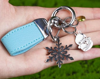 May include: A keychain with a light blue leather strap and silver-tone metal hardware. The keychain features a snowflake charm and a snowman charm. The background is a green, textured surface.