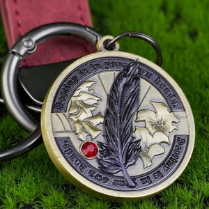 May include: A circular, antique-style pendant keychain with the words "Destiny is in your own hands." The pendant displays a feather, maple leaves, and a red jewel. The keychain has a silver clasp and a burgundy leather strap.
