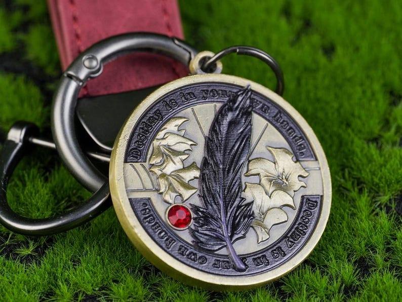 May include: A circular, antique-style pendant keychain with the words "Destiny is in your own hands." The pendant displays a feather, maple leaves, and a red jewel. The keychain has a silver clasp and a burgundy leather strap.