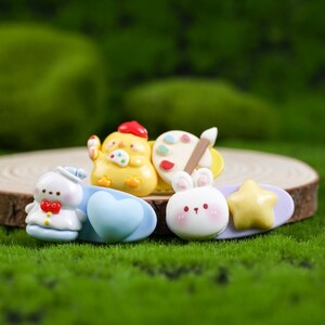 Peut inclure: Ensemble de barrettes à cheveux colorées de style dessin animé. Les clips présentent divers motifs, dont un poussin jaune avec un pinceau, un lapin blanc, une étoile et un personnage avec un cœur. Ils sont posés sur une tranche de bois sur fond vert.