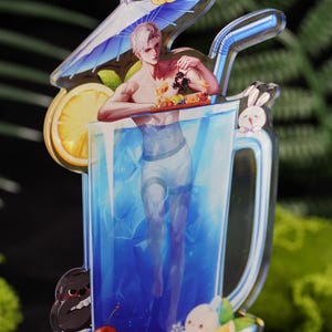 May include: A clear acrylic charm in the shape of a large glass of blue liquid with a straw. The charm features a shirtless man standing in the glass, holding a tray of food. A lemon slice, a small umbrella, and cartoon characters are also included.