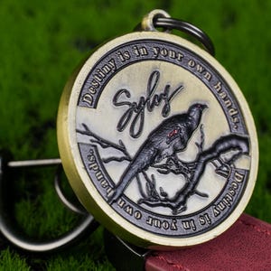 May include: A circular, antique-style keyring featuring a bird on a branch. The keyring is inscribed with the words "Destiny is in your own hands" and the name "Sylas". The bird is black and grey, and the keyring has a gold and silver finish.