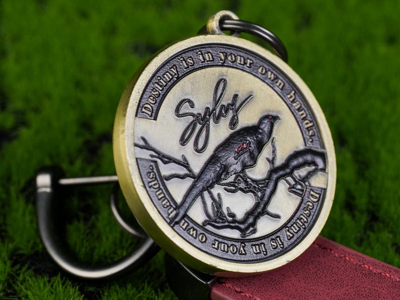 May include: A circular, antique-style keyring featuring a bird on a branch. The keyring is inscribed with the words "Destiny is in your own hands" and the name "Sylas". The bird is black and grey, and the keyring has a gold and silver finish.