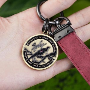 May include: Keychain with a claret leather strap and a circular, bronze-coloured pendant. The pendant features a bird perched on a branch, the word "Syby", and the text "Destiny is in your own hands."