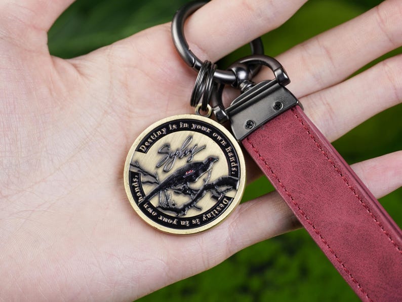 May include: Keychain with a claret leather strap and a circular, bronze-coloured pendant. The pendant features a bird perched on a branch, the word "Syby", and the text "Destiny is in your own hands."