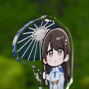 May include: A clear acrylic charm featuring a cartoon character holding a parasol. The parasol has a blue and white design. The character has brown hair and is wearing a white and blue outfit. The background is a blurred green.