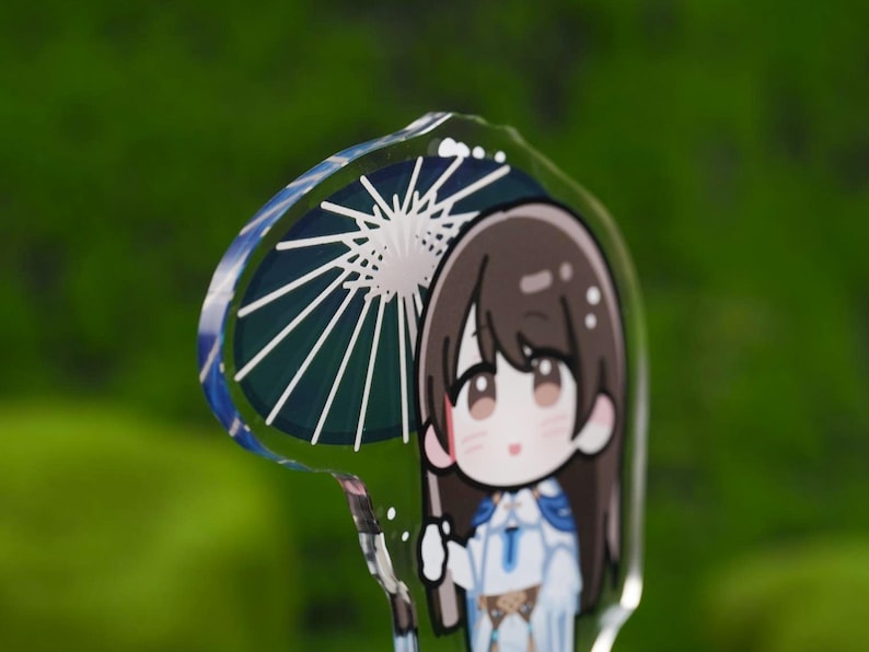 May include: A clear acrylic charm featuring a cartoon character holding a parasol. The parasol has a blue and white design. The character has brown hair and is wearing a white and blue outfit. The background is a blurred green.
