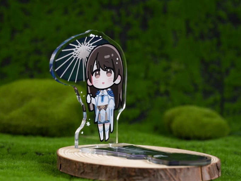 May include: A clear acrylic stand featuring a cartoon character with long brown hair, holding a parasol. The character is wearing a white and blue outfit. The stand is on a wooden base, set against a green, mossy background.