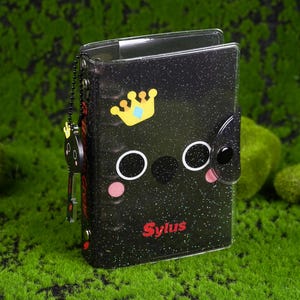 Puede incluir: Cuaderno negro brillante con un diseño de cara de dibujos animados, con una corona, ojos delineados en blanco y mejillas rosadas. La palabra "Sylus" está escrita en rojo. Un pequeño dije con una llave cuelga de una cadena.