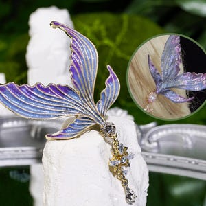 Peut inclure: Une barrette en forme de queue de sirène avec une finition en métal doré. La queue présente un dégradé d'émail bleu et violet avec un charm en cristal pendant. Un encart circulaire montre la barrette en utilisation.