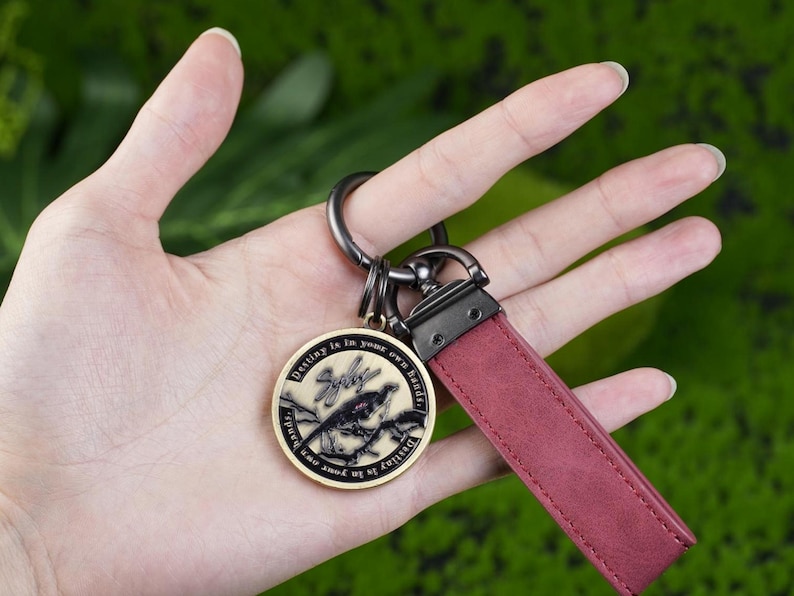 May include: A keychain with a claret leather strap and a round, gold-coloured pendant. The pendant features a bird design and the text "Destiny is in your own hands". The keychain has a metal ring and clasp.