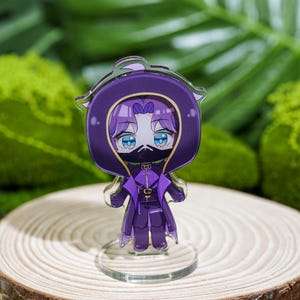 Peut inclure: Figurine acrylique violette de style anime repr&eacute;sentant un personnage avec une cape &agrave; capuche et un masque. La figurine a les yeux bleus et se tient sur une base transparente, plac&eacute;e sur une tranche de bois. Le fond pr&eacute;sente un feuillage vert.