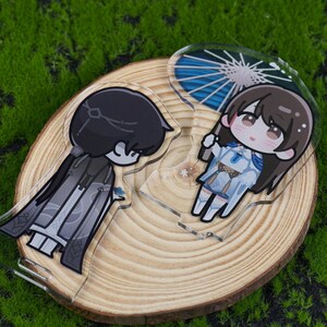 May include: Two acrylic anime-style character charms. One charm features a dark-haired character in a black and gray outfit. The other charm depicts a character with brown hair and a blue and white outfit, holding a blue parasol. Both charms are on a wooden slice.
