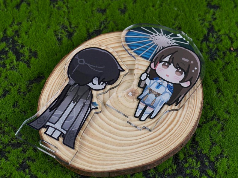 May include: Two acrylic anime-style character charms. One charm features a dark-haired character in a black and gray outfit. The other charm depicts a character with brown hair and a blue and white outfit, holding a blue parasol. Both charms are on a wooden slice.