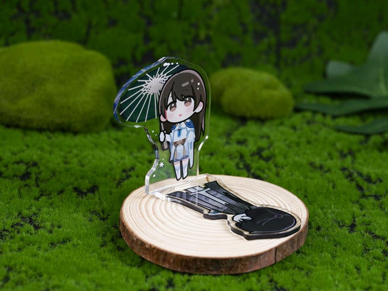 May include: A clear acrylic charm featuring a cartoon character holding an umbrella. The charm is on a wooden disc with a black and white design. The background is a green mossy surface.